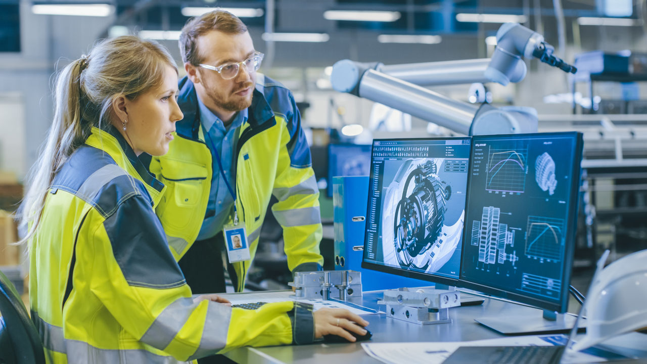 At the Factory: Male Mechanical Engineer and Female Chief Engineer Work Together on the Personal Computer, They Discuss Details of the 3D Engine Model for Robotic Arm.